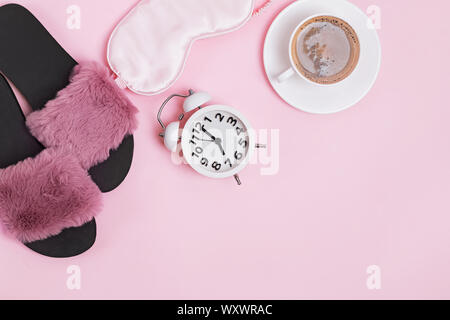 Accogliente mattina a casa. Caffè, mascherina, pantofole e sveglia su sfondo rosa con spazio di copia Foto Stock