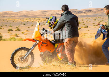 Merzouga, Marocco - 25 Febbraio 2016: una moto rider agita la sabbia come egli tenta di ottenere la sua bici arancione di andare nel deserto del Sahara. Due uomini sono com Foto Stock