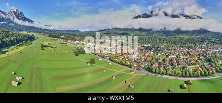 Panorama da composita le fotografie aeree di una casa vacanze insediamento dietro un prato con dei fienili e stalle di fronte alle enormi scenario delle Alpi. Foto Stock