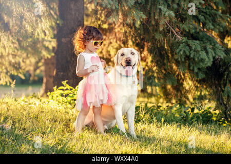 Carino adorabili poco ricci ragazza caucasica indossando occhiali da sole giallo con il suo cane nel parco al di fuori al tramonto sul giorno di estate. Bambini che giocano con sottoprodotti di origine animale Foto Stock