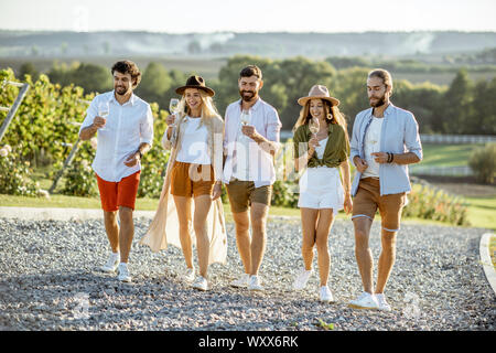 Un gruppo di giovani amici vestito casualmente appendere fuori insieme, camminando con bicchieri di vino sul vigneto in una giornata di sole Foto Stock
