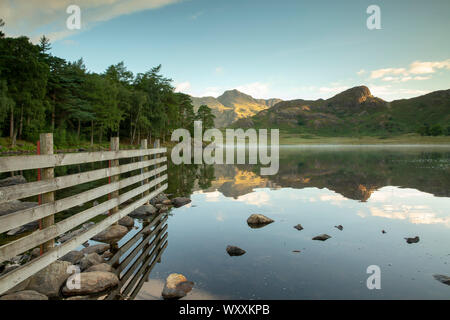 Blea Tarn Parco Nazionale del Distretto dei Laghi con The Langdale Pikes nella distanza riflettendo sul lago durante un sunrise e qualche foschia sulla superficie di Foto Stock