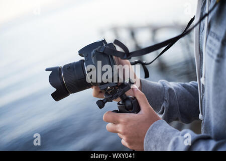 Un uomo mette la fotocamera reflex sulla testa di un treppiede per scattare foto da parte del mare o di un lago con acqua blu e un molo di legno in background Foto Stock