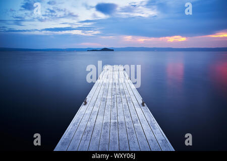 Un vecchio molo in legno per le barche sul lago di Bolsena a Capodimonte, sull'acqua, di fronte all'Isola Bisentina, durante il tramonto con nubi colorate e su Foto Stock